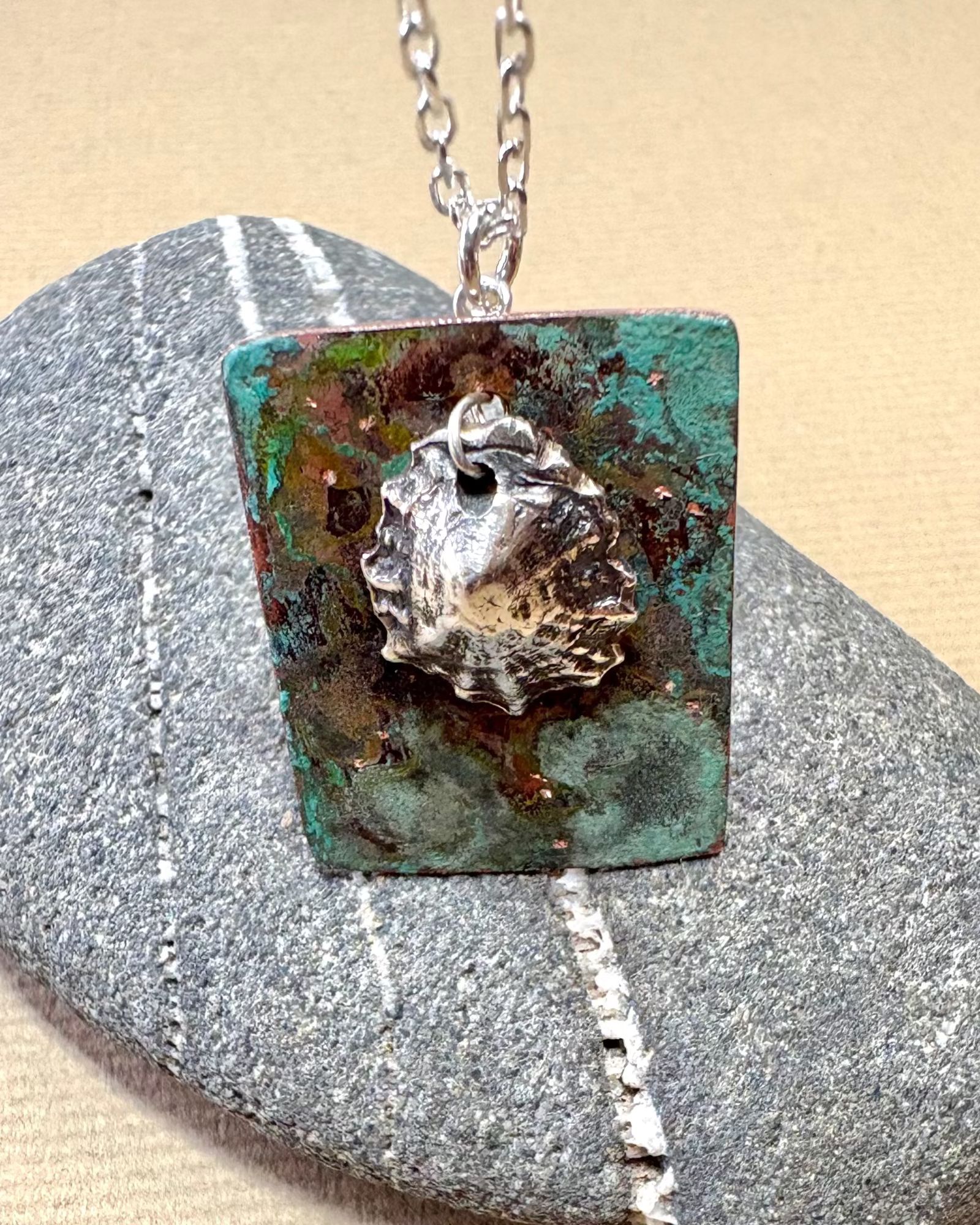 Silver limpet shell in its rock pool with a square pendant featuring an oxidised pattern on copper on a pebble surface