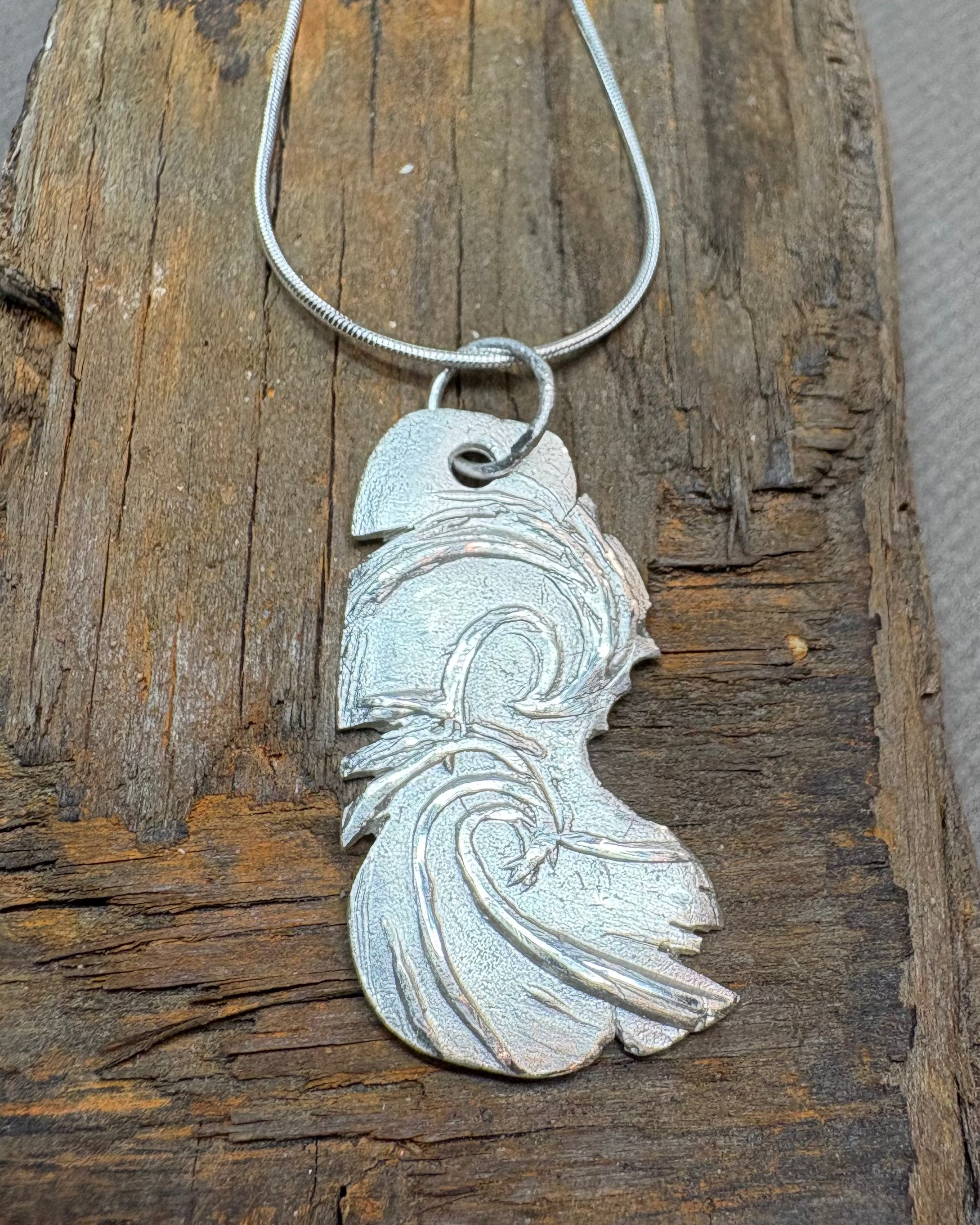 Silver surf pendant on a silver cord chain, placed on a piece of driftwood