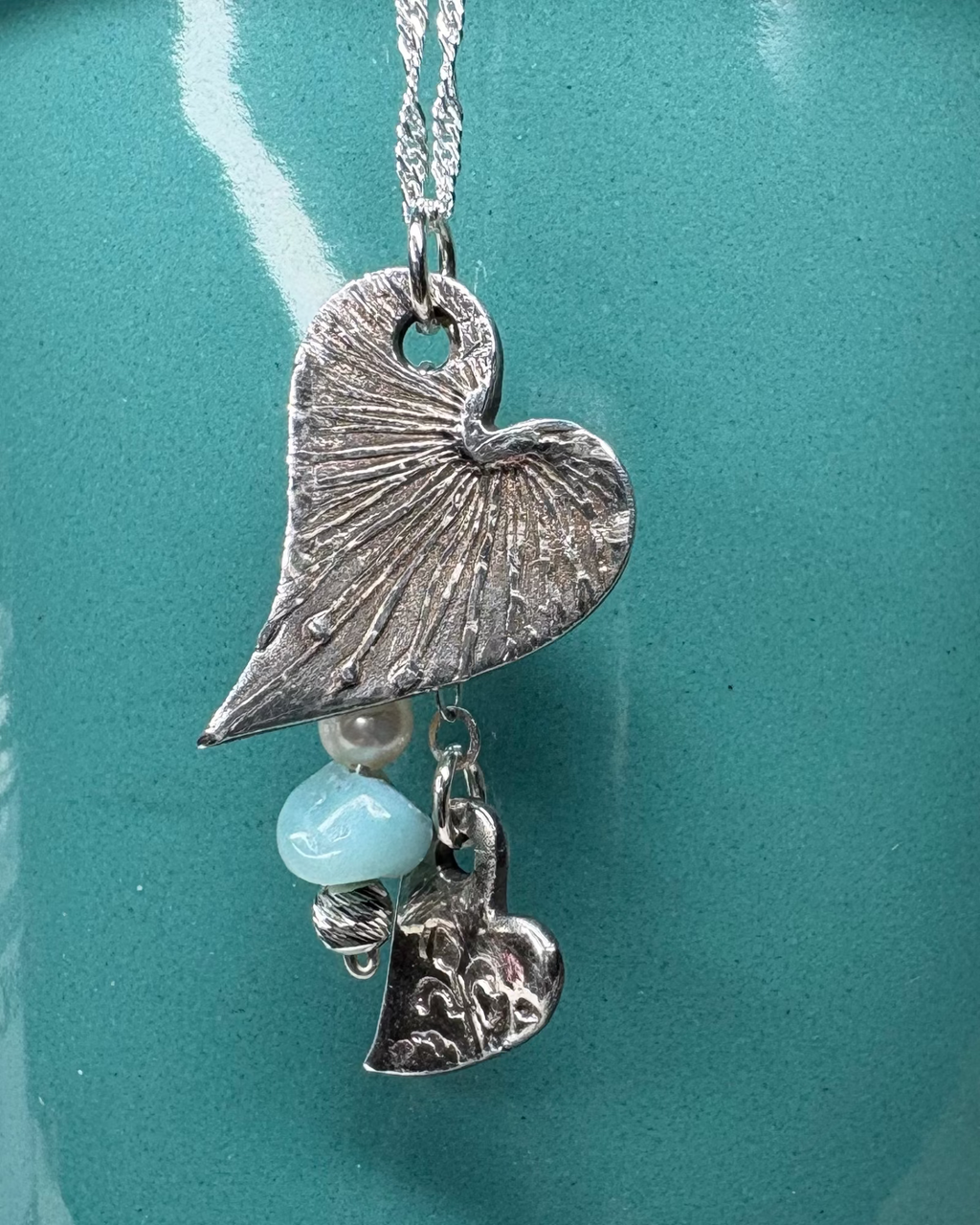Necklace with 2 heart-shaped charms with pearl and apatite bead with silver chain on a teal background.