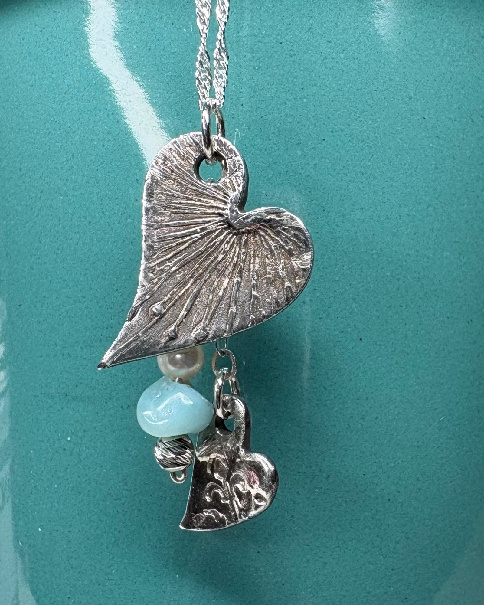 Necklace with 2 heart-shaped charms with pearl and apatite bead with silver chain on a teal background.