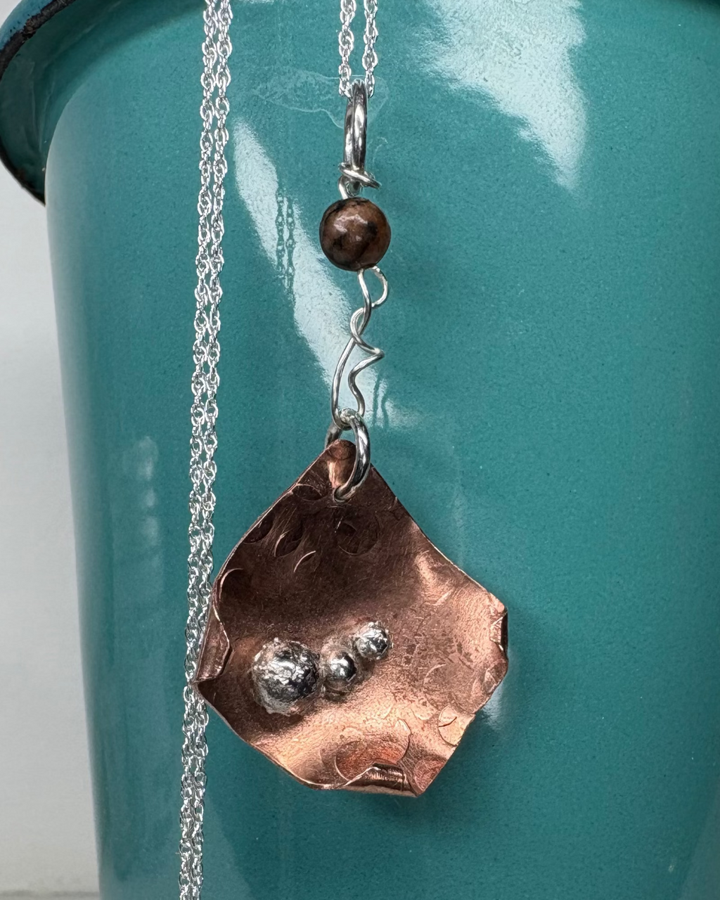 Textured copper necklace with curled corner design and silver orbs with a silver chain against a teal background