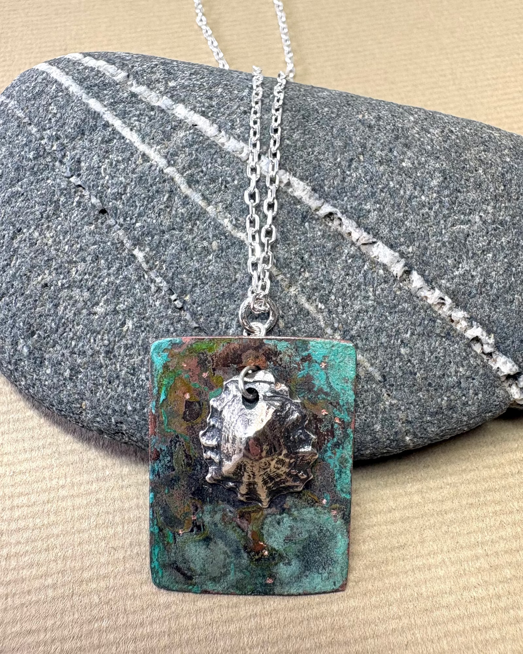 Silver limpet shell in its rock pool with a square pendant featuring a natural pattern  on copper on a pebble surface