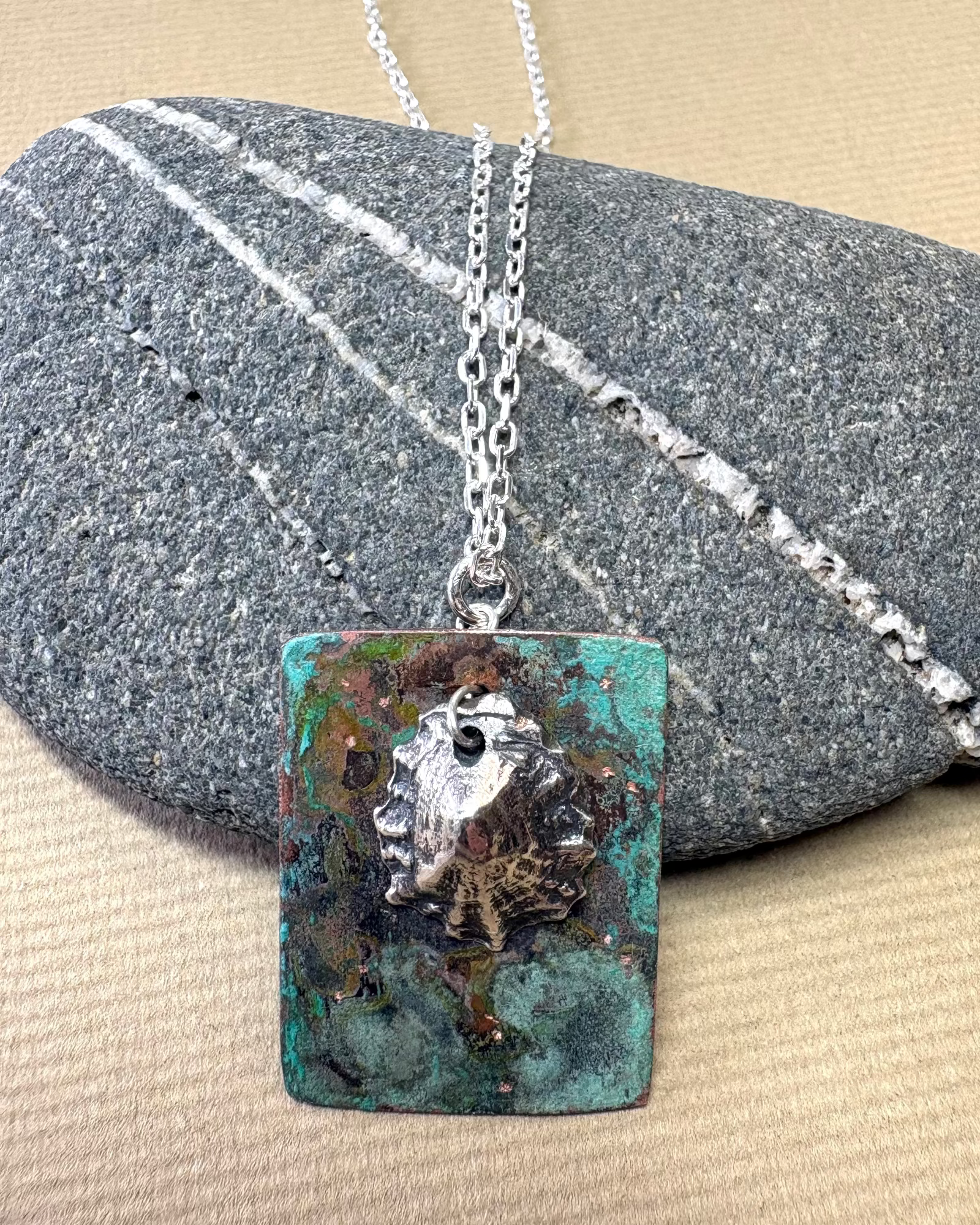 Silver limpet shell in its rock pool with a square pendant featuring a natural pattern  on copper on a pebble surface