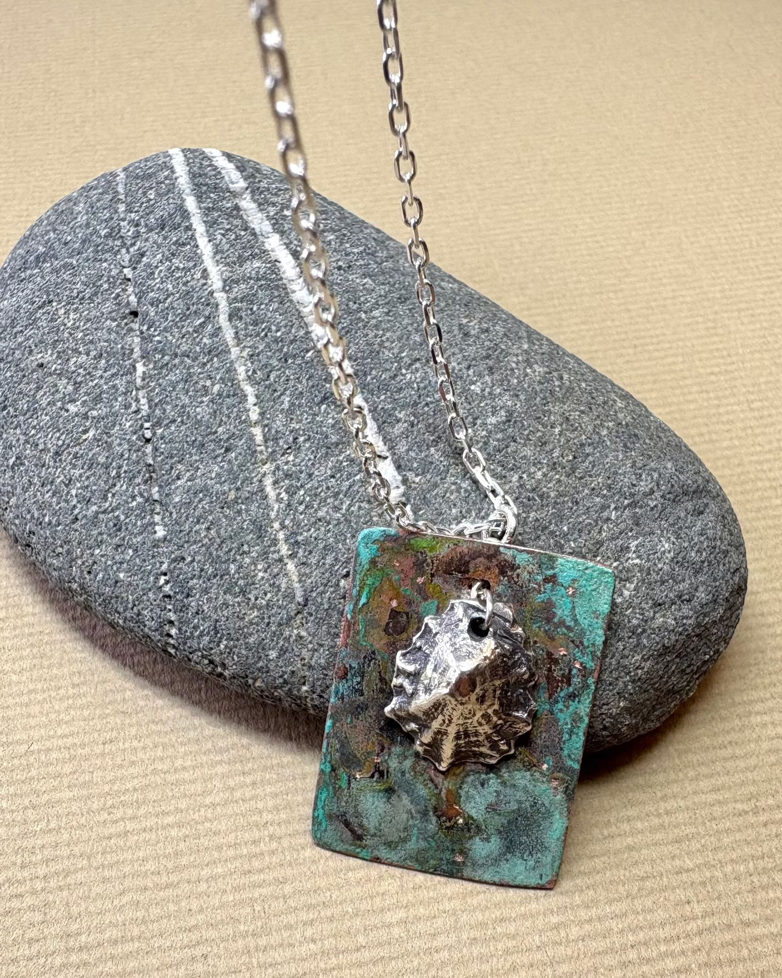 Silver limpet shell in its rock pool with a square pendant featuring an oxidised pattern on copper on a pebble surface