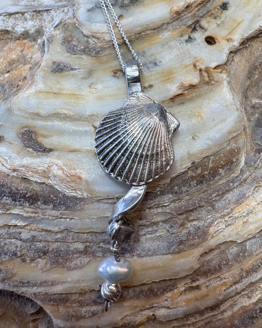 Silver scallop shell necklace with a pearl pendant on a textured shell surface