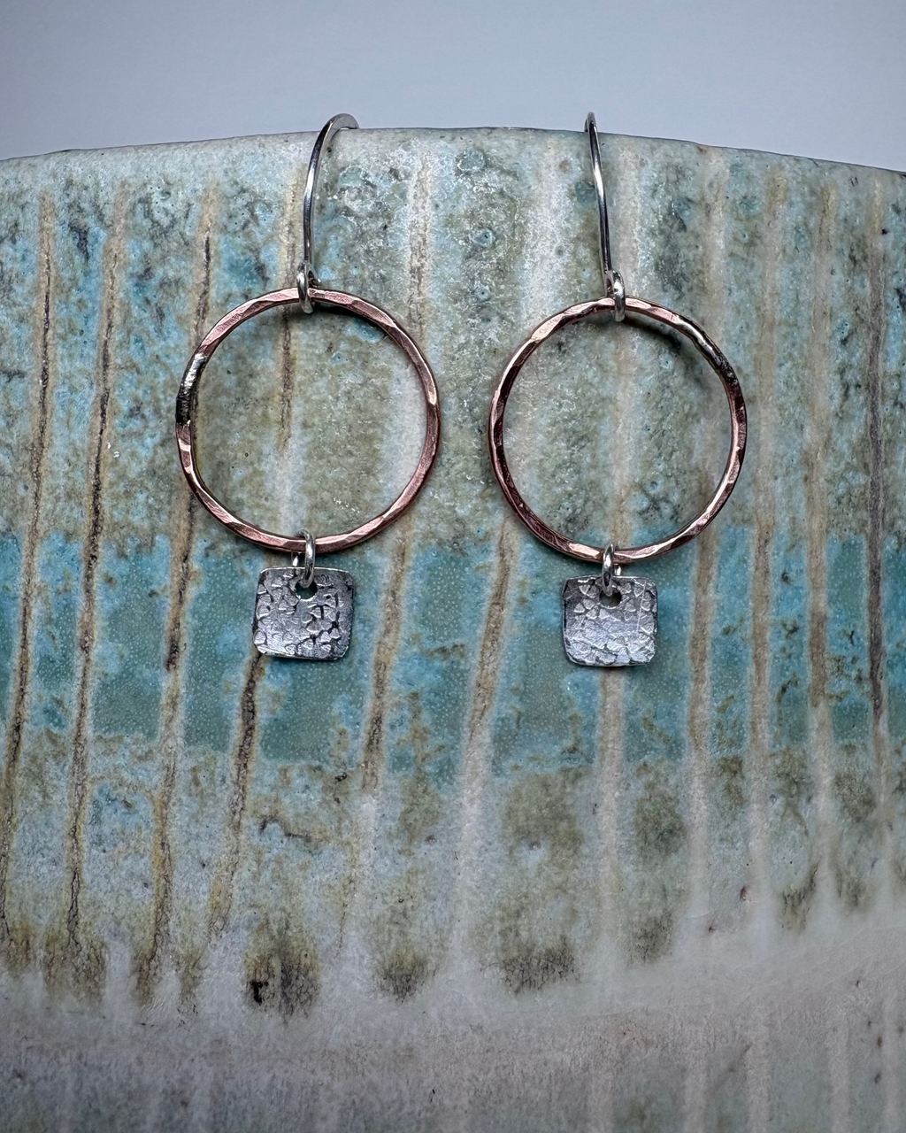 Pair of bronze hoop earrings with square silver charms on a ceramic surface.