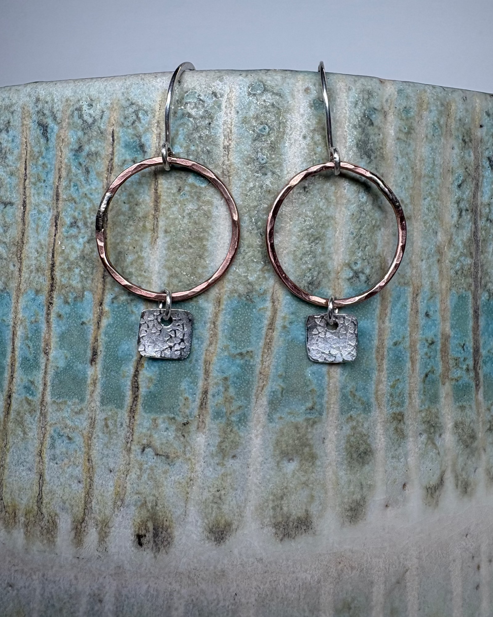 Pair of bronze hoop earrings with square silver charms on a ceramic surface.