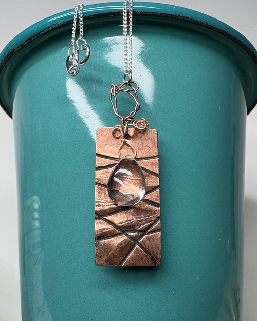 Copper rectangle textured necklace on a silver chain with wired teardrop quartz over a teal container.