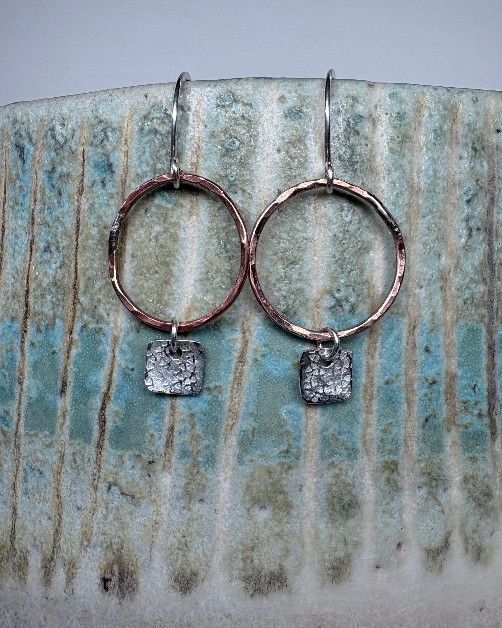 Pair of bronze hoop earrings with silver textured square charms on a ceramic surface.