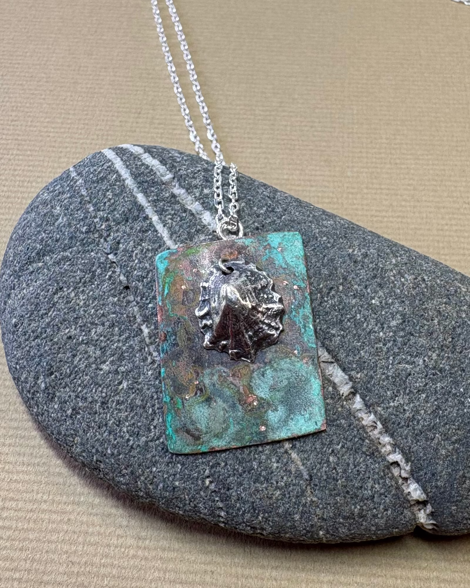 Silver limpet shell in its rock pool with a square pendant featuring an oxidised pattern on copper on a pebble surface