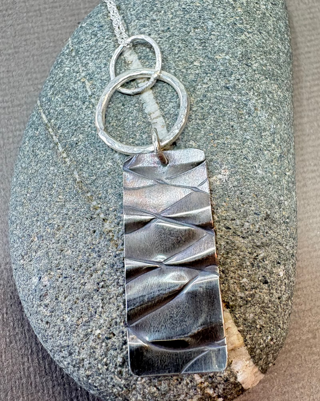 Silver hoops and textured rectangle pendant on a pebble surface