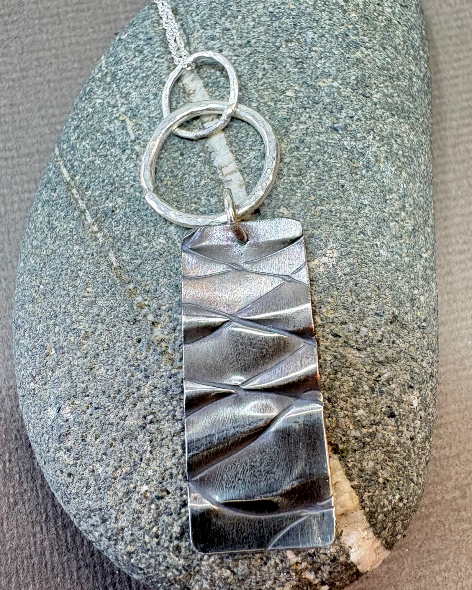 Silver hoops and textured rectangle pendant on a pebble surface