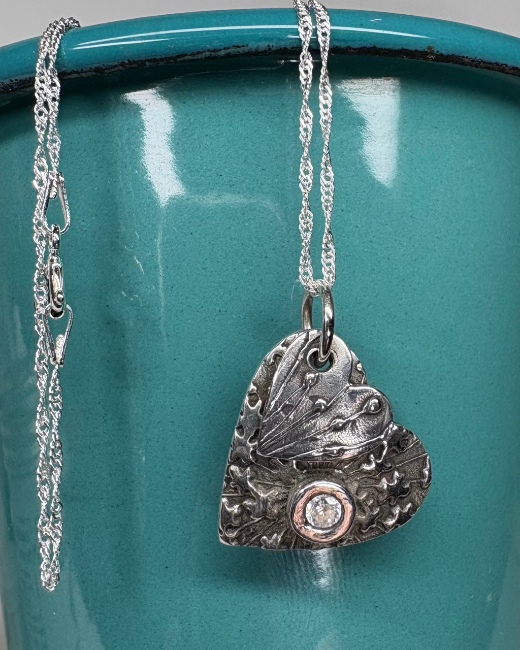 Silver oxidised double heart necklace with textured seed head design hanging on a silver chain pictured on a teal background