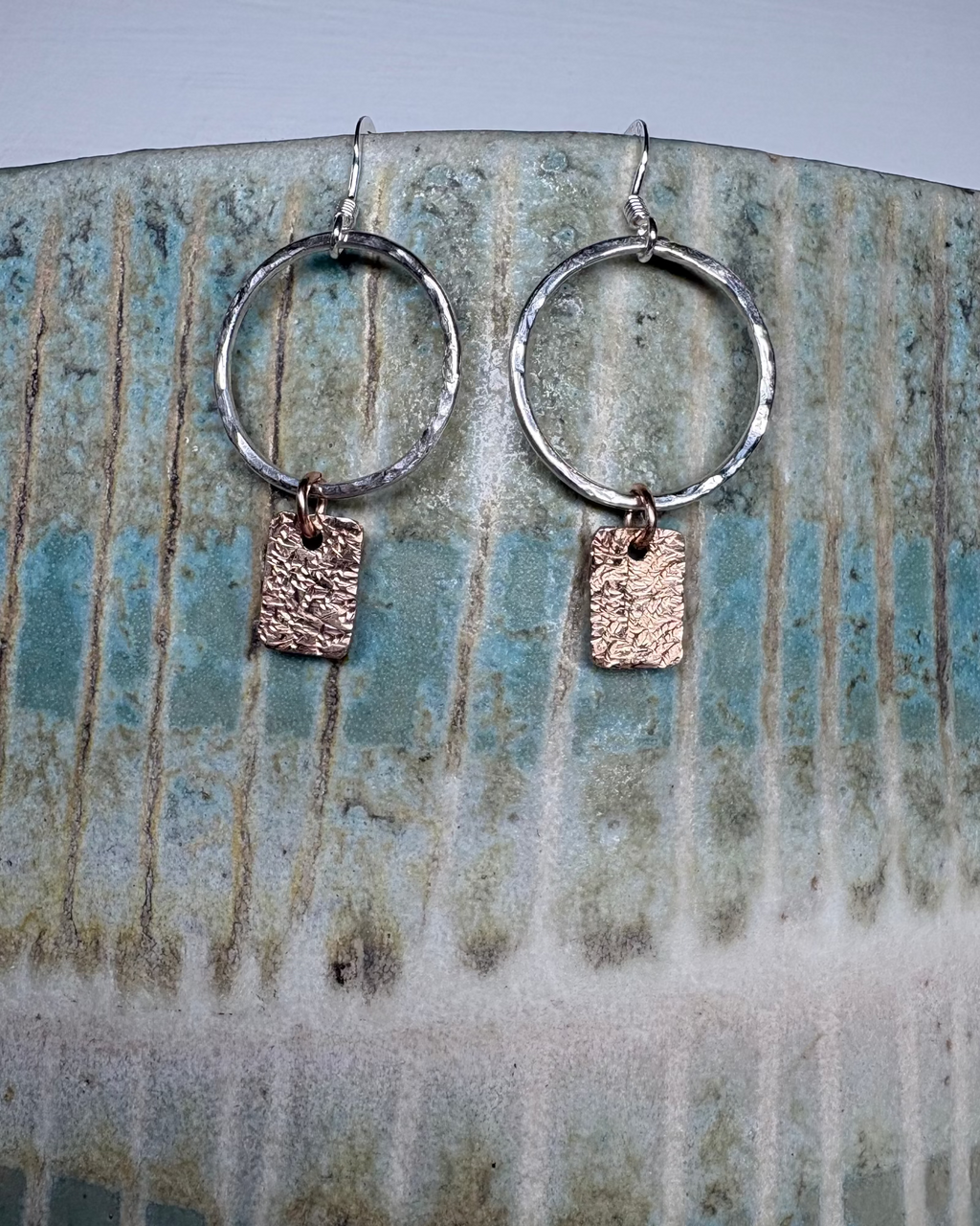 Pair of hammered silver hoop earrings with bronze rectangular charms on a ceramic surface.