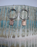 Pair of hammered silver hoop earrings with bronze rectangular charms on a ceramic surface.