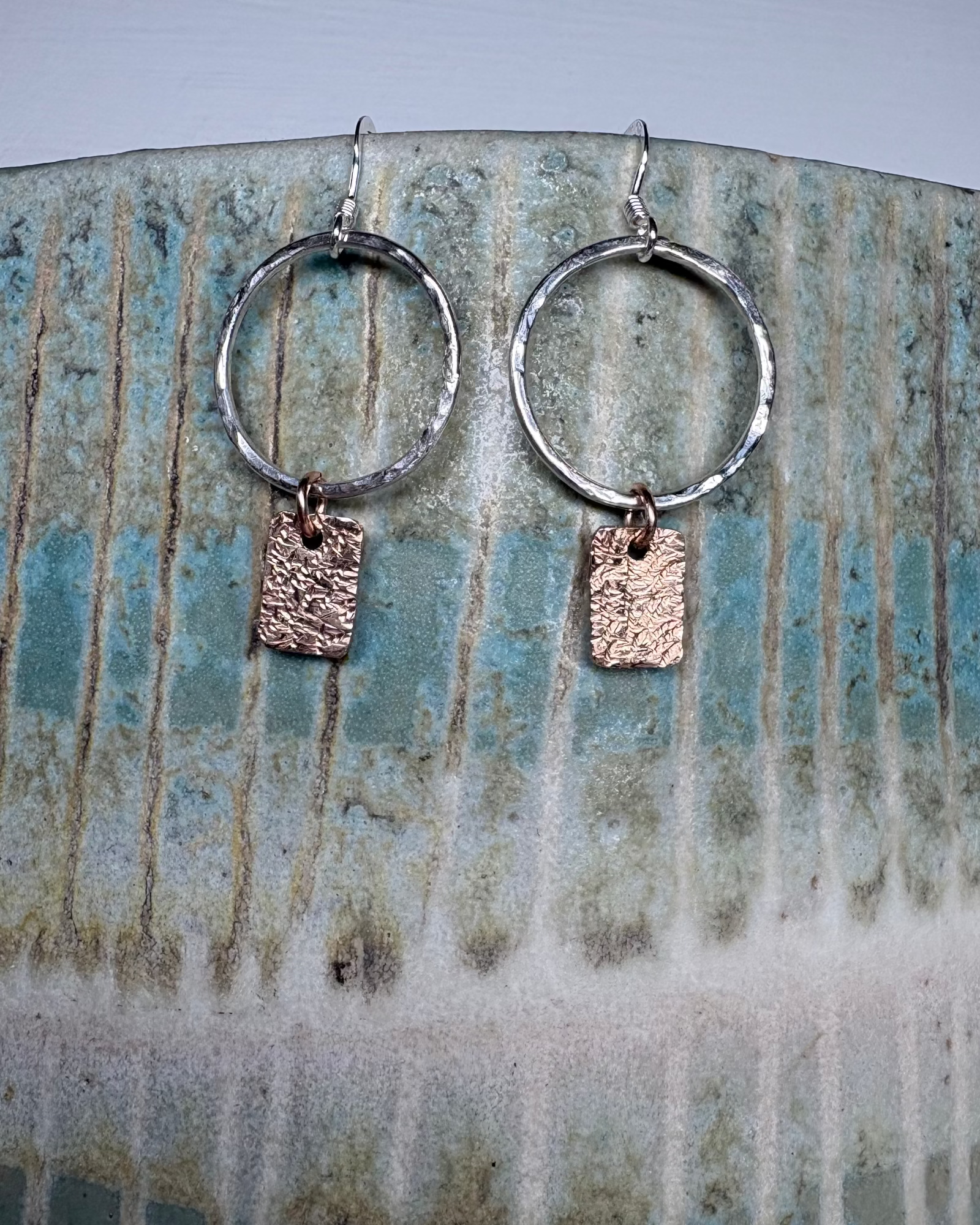 Pair of hammered silver hoop earrings with bronze rectangular charms on a ceramic surface.