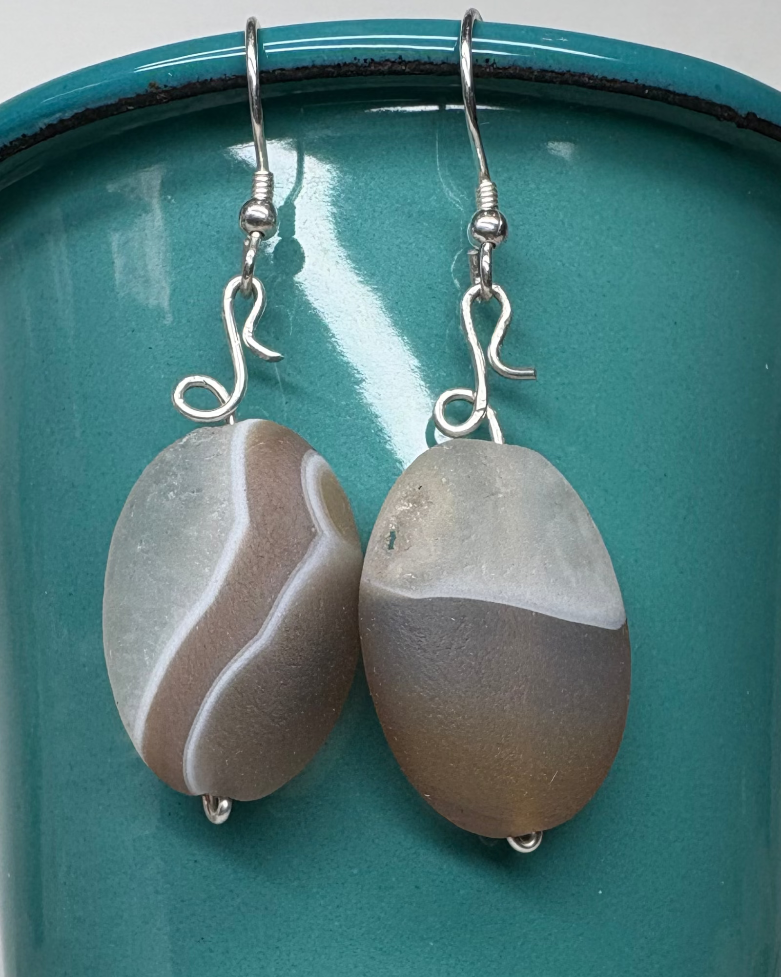 Pair of earrings with matt agate geode ovals wired with silver pictured on a teal cup
