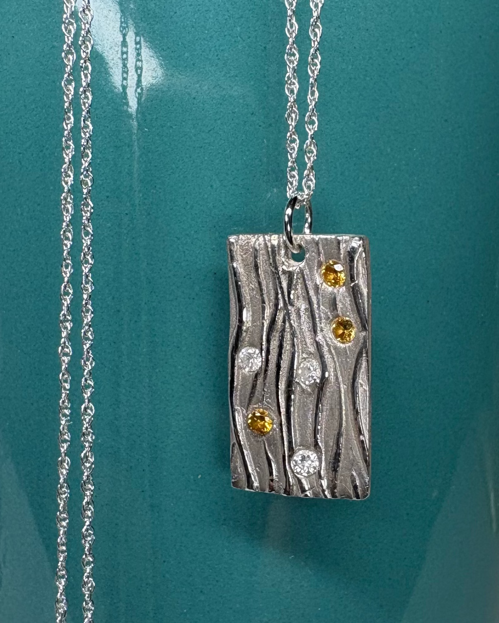 Silver rectangle necklace with wavy design and white and gold zirconia a silver chain on a teal cup