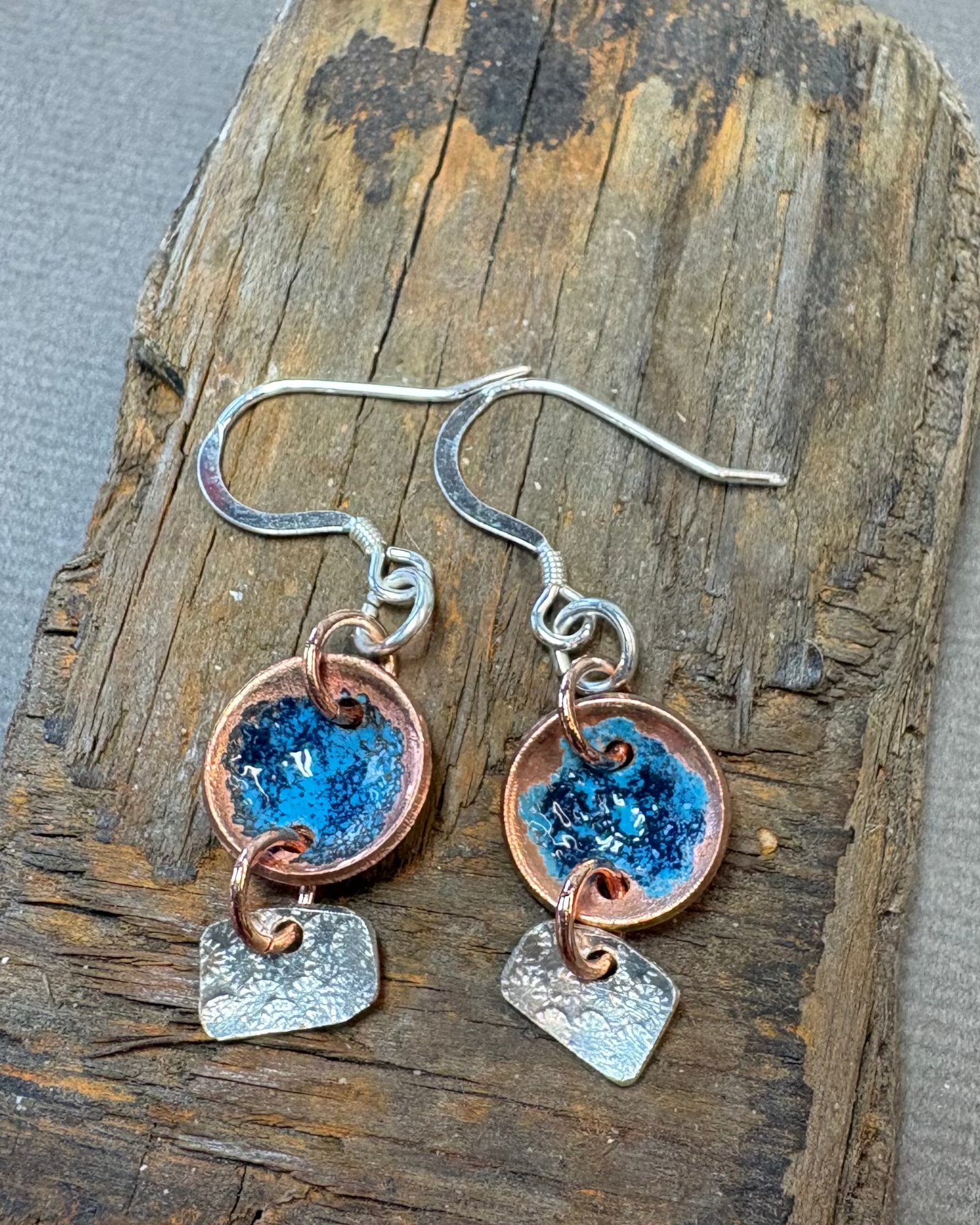 Pair of copper earrings with blue enamel with silver hanging texture shapes on a piece of driftwood