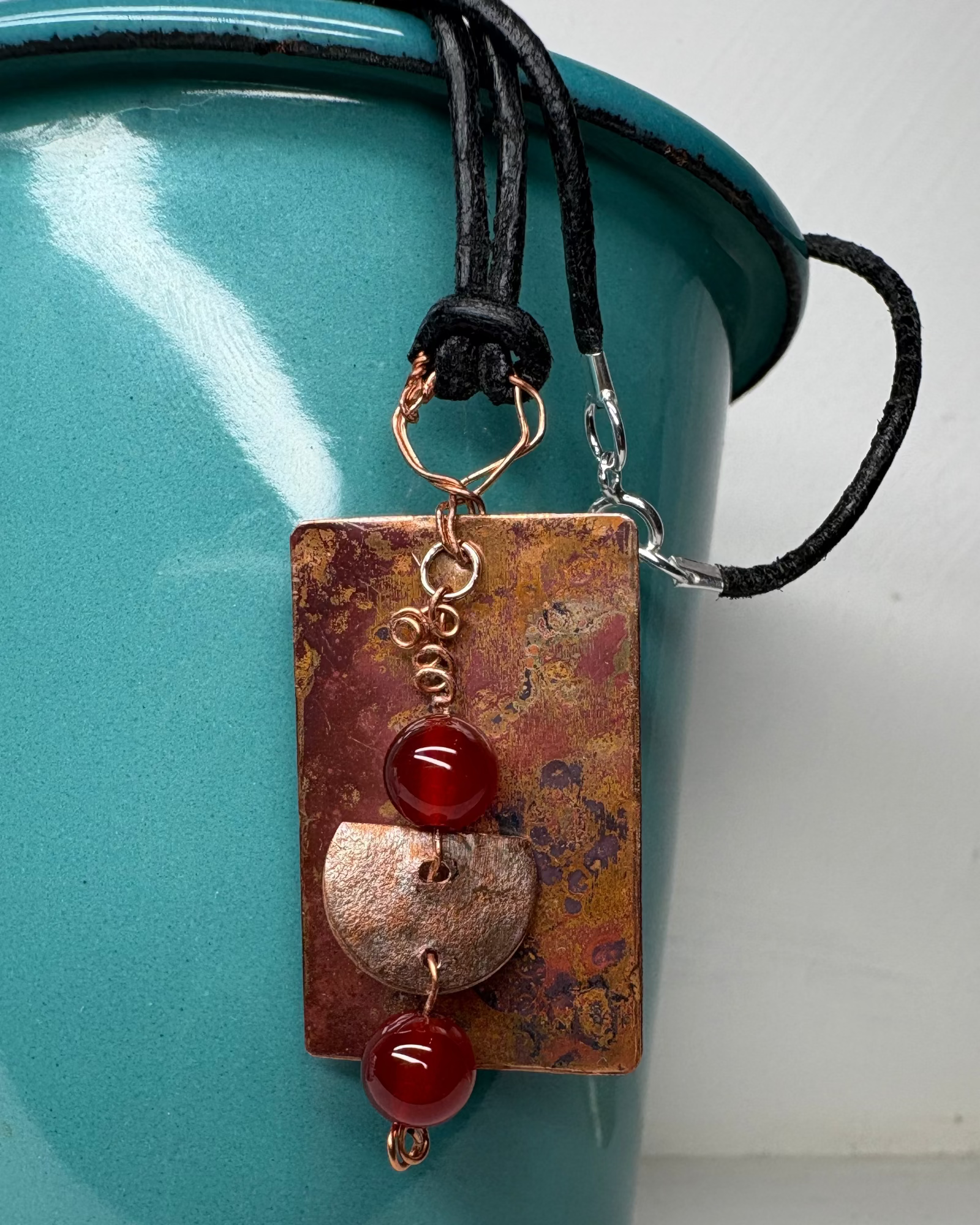 Copper rectangular oxidised patina pendant featuring red carnelian stones and copper half moon hanging charm with a black leather cord against a teal container.