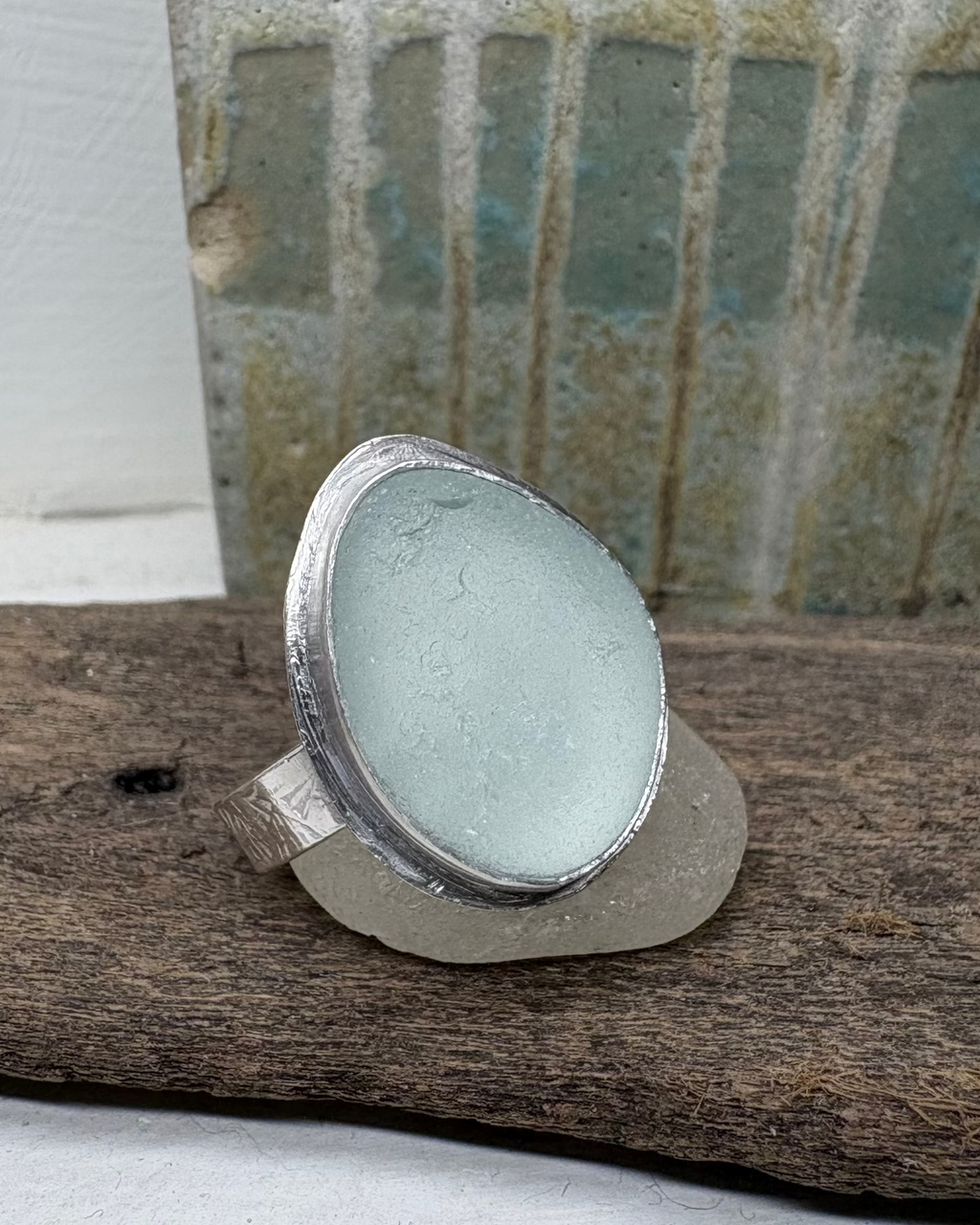 Silver halo set ring with a light aqua sea glass on a silver band shown on a piece of sea glass on driftwood