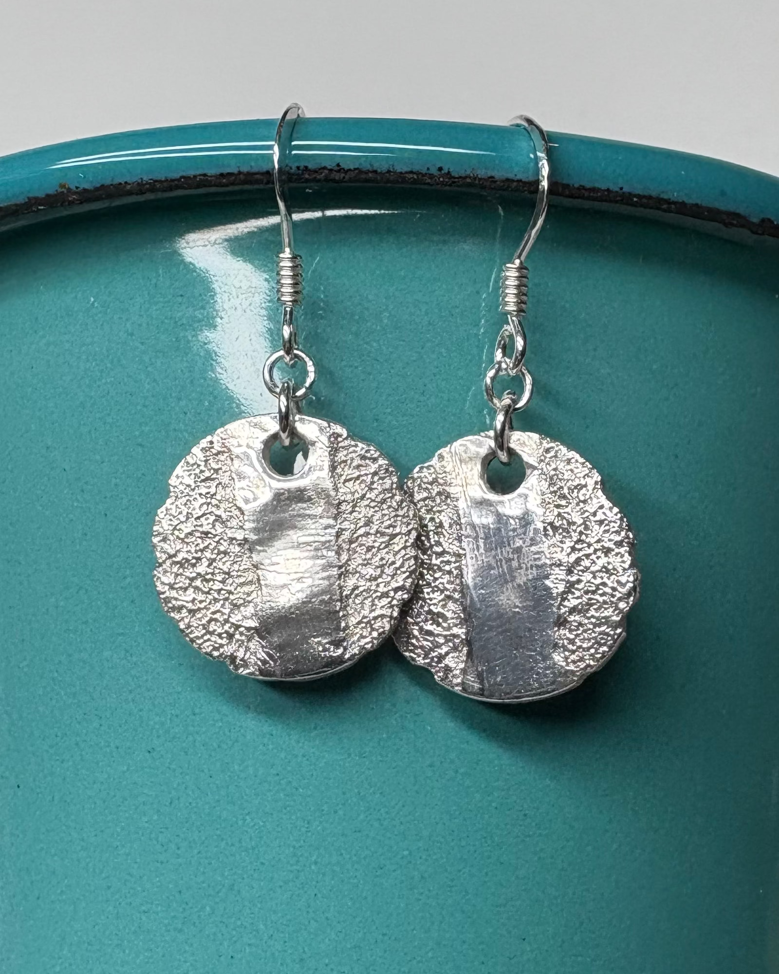 Silver textured and polished circular earrings hanging from a teal container 
