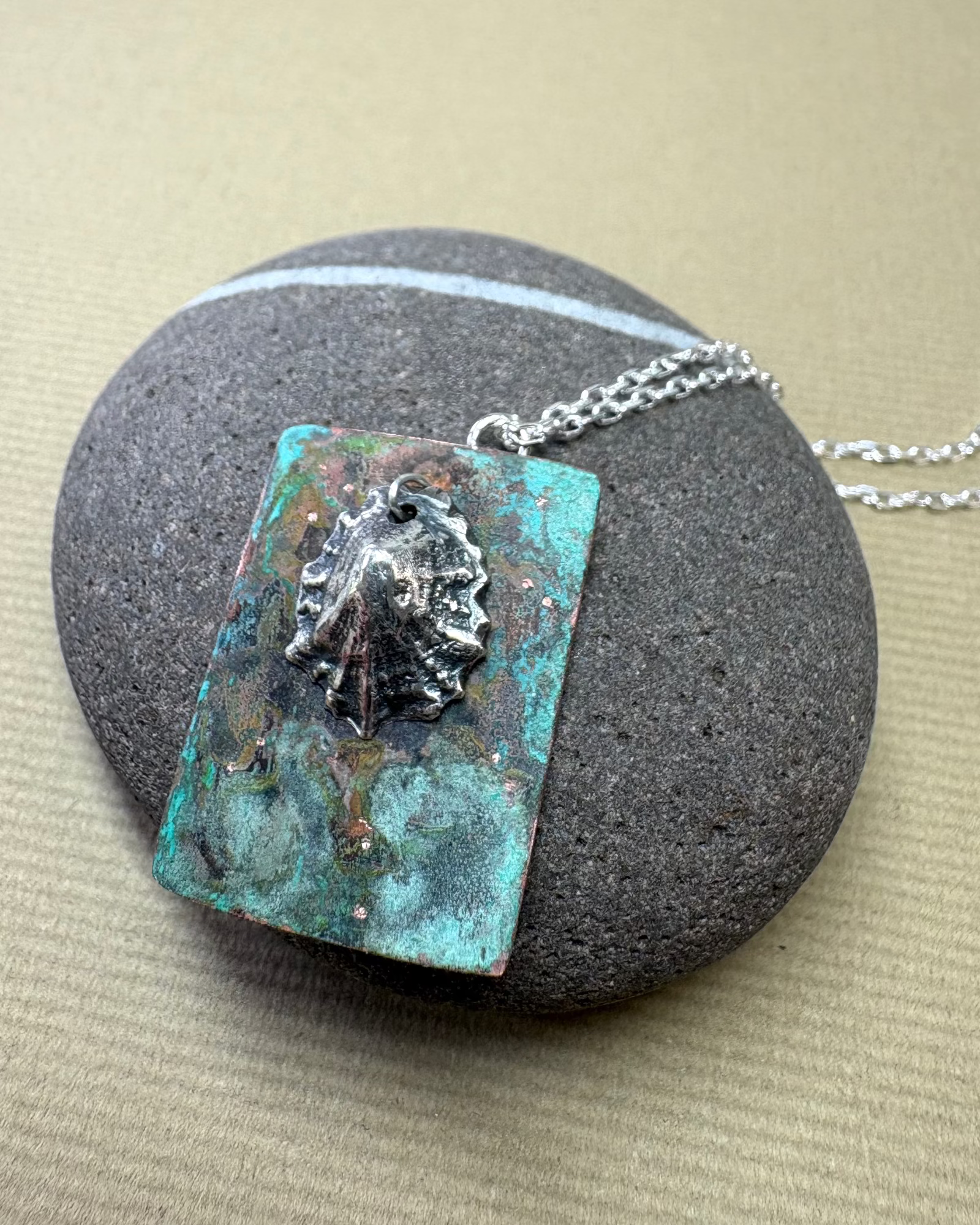 Silver limpet shell in its rock pool with a square pendant featuring an oxidised pattern on copper on a pebble surface