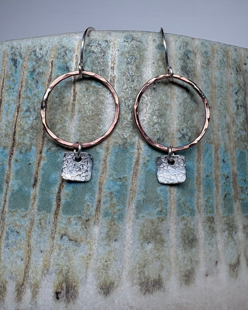 Bronze hoop earrings with textured square silver charms on a ceramic surface.