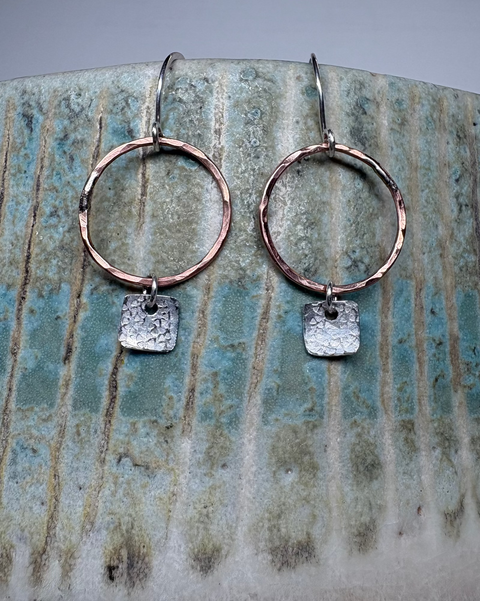 Bronze hoop earrings with textured square silver charms on a ceramic surface.