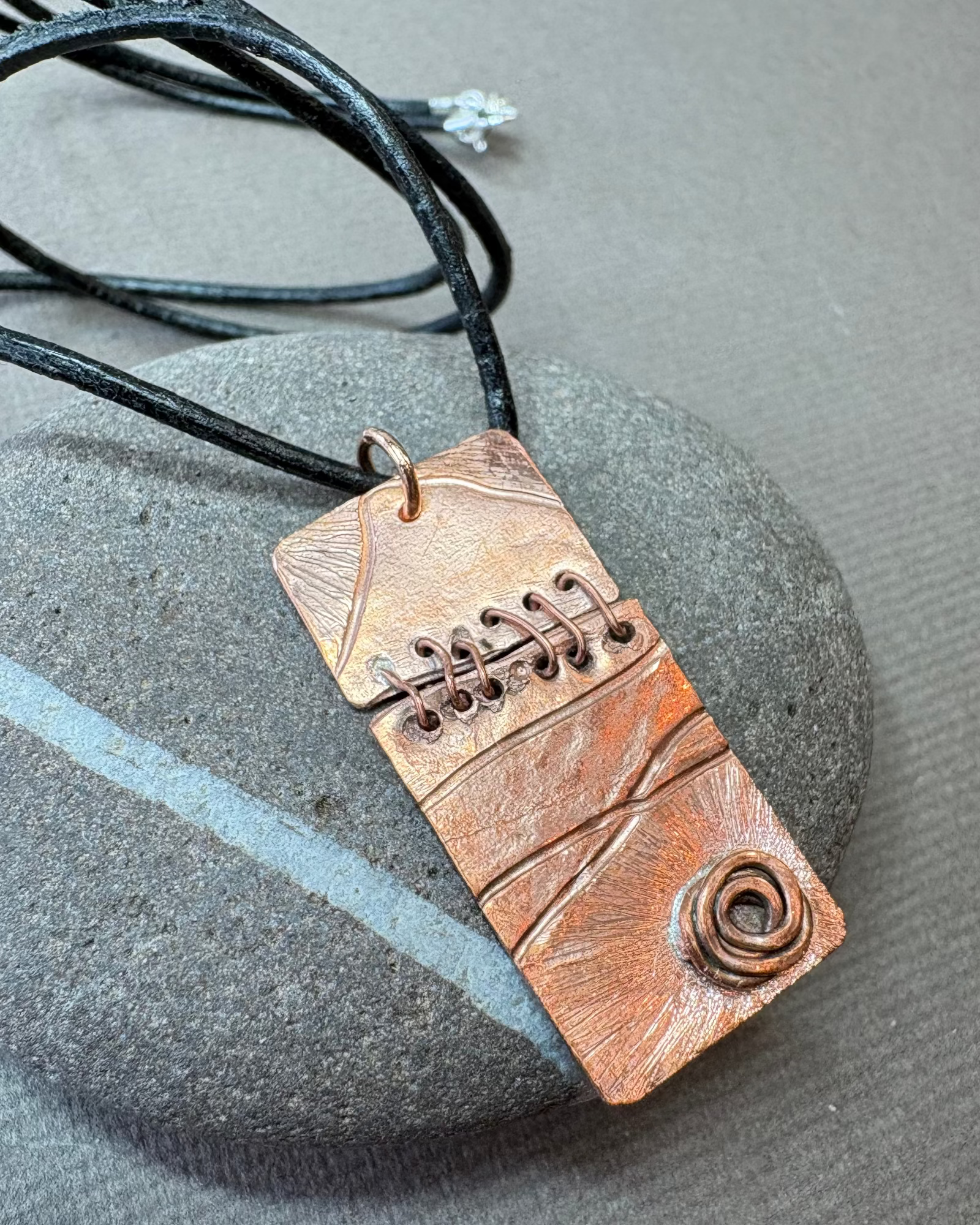 Copper stitched texture pendant necklace with black leather cord on a pebble