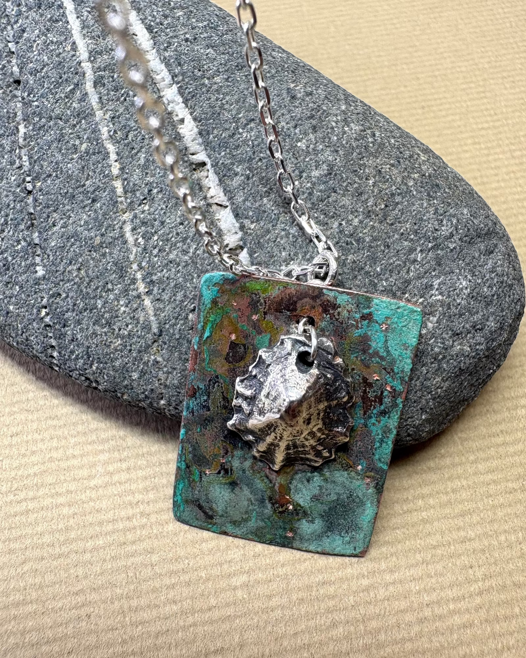 Silver limpet shell in its rock pool with a square pendant featuring an oxidised pattern  on copper on a pebble surface