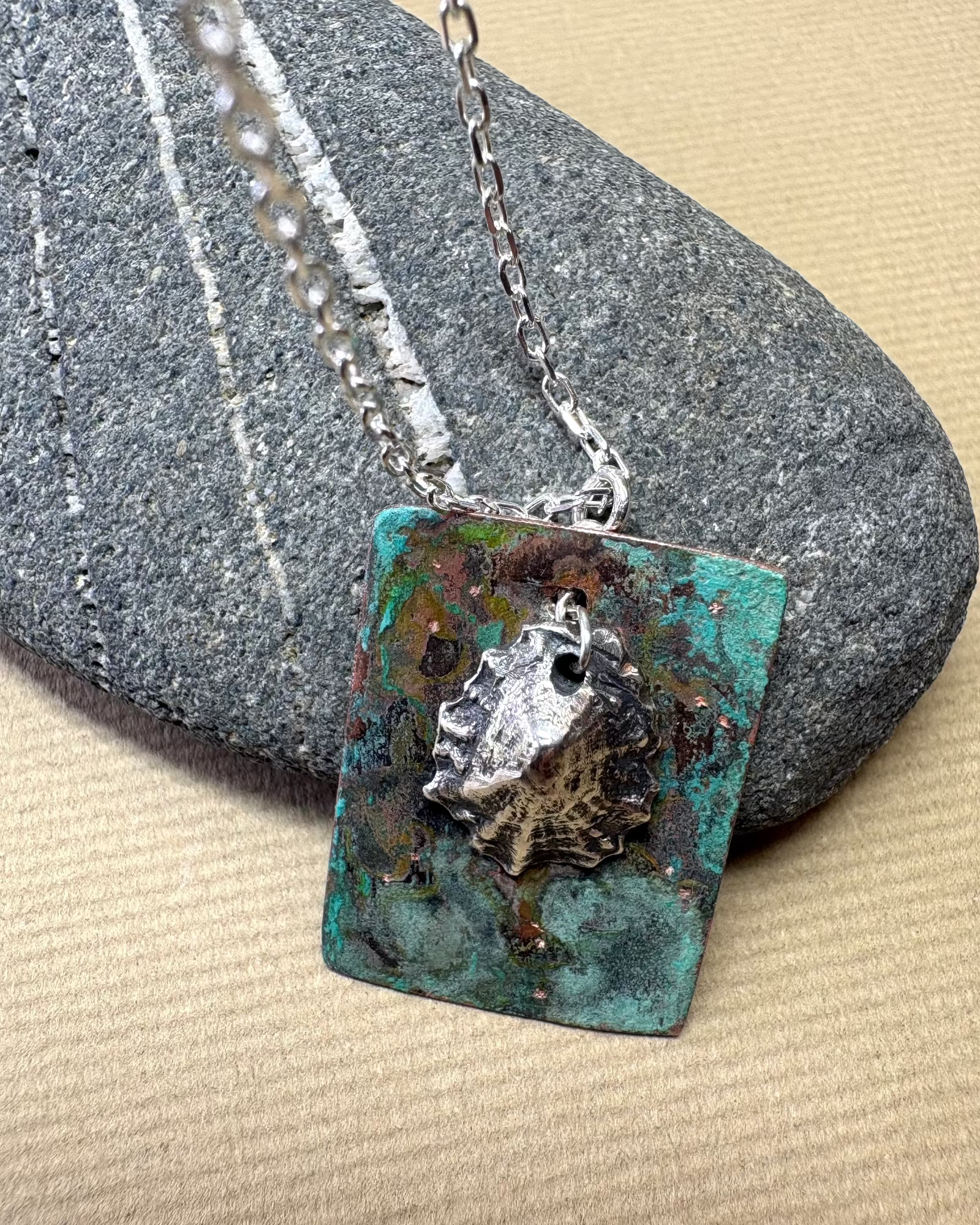Silver limpet shell in its rock pool with a square pendant featuring an oxidised pattern  on copper on a pebble surface