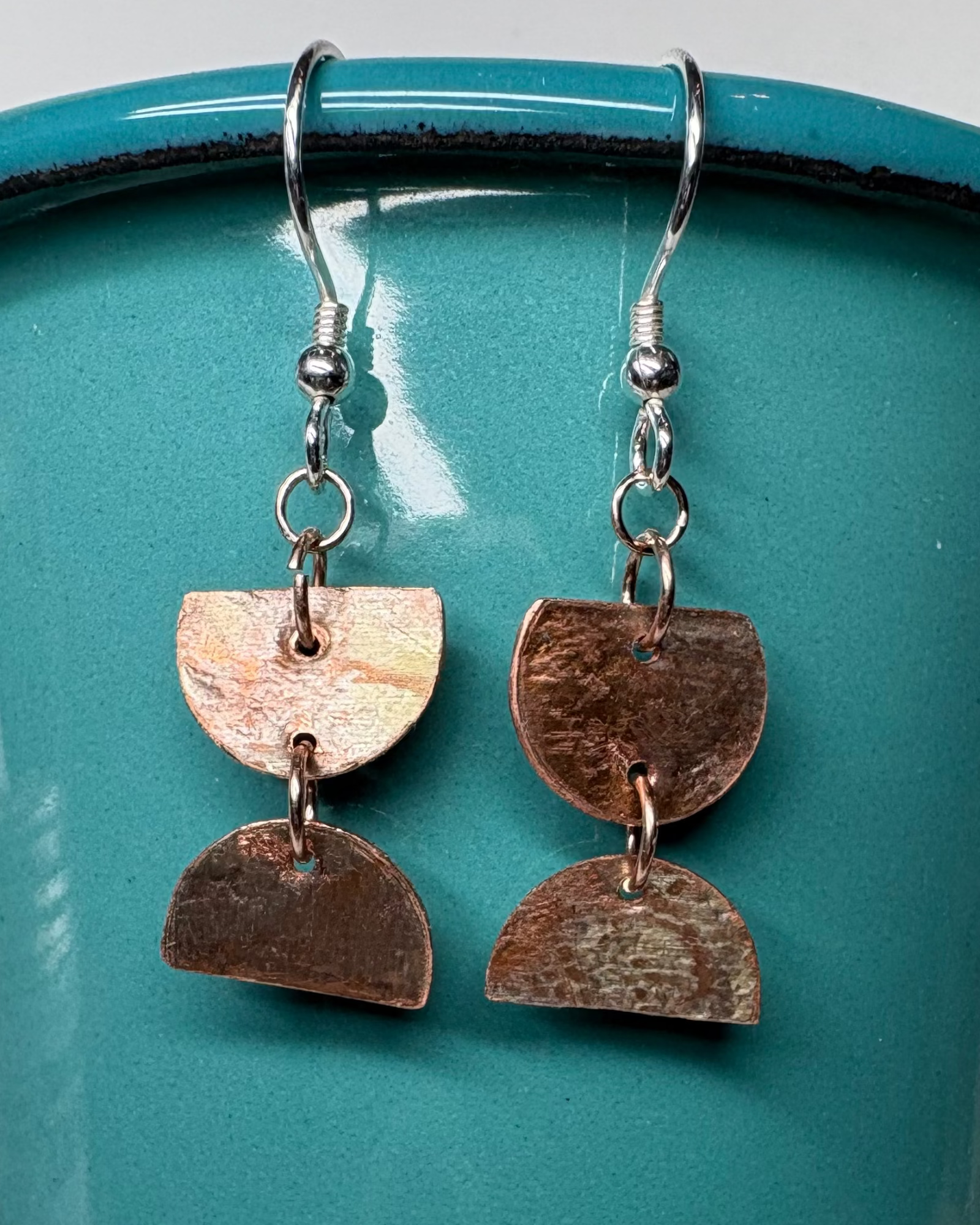 Pair of textured circular cut earrings with patina hanging from a teal cup 