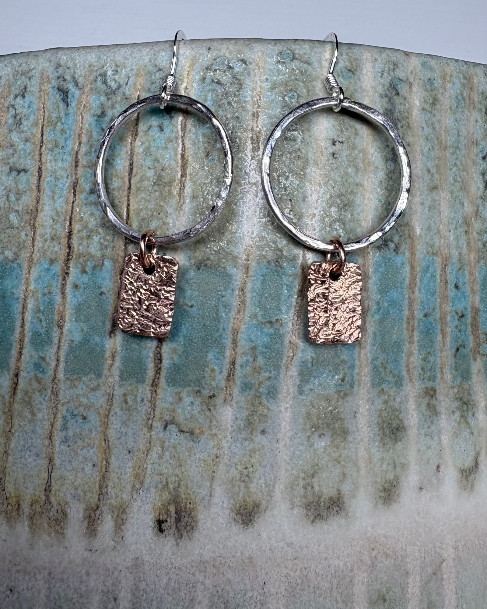Silver hoop earrings with bronze rectangular charms on a ceramic plate.