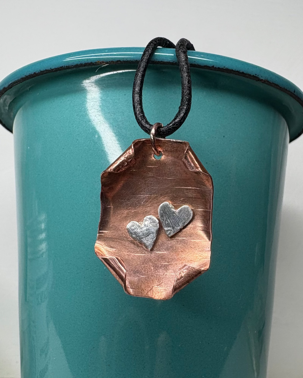 Rectangular copper necklace with curled corner design with two fine silver hearts with black leather cord pictured on a teal cup