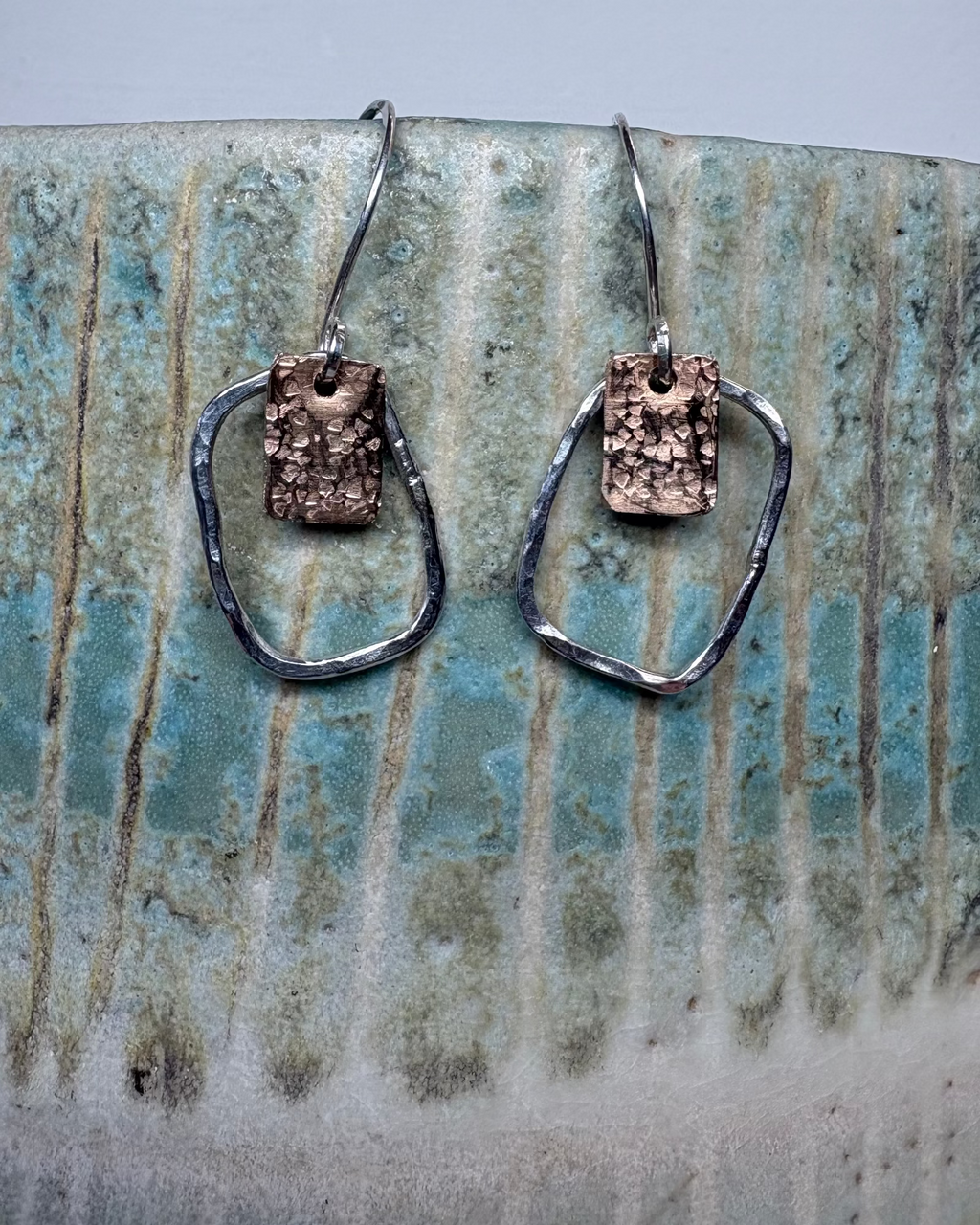 Pair of silver hoop earrings with bronze textured squares on a ceramic surface