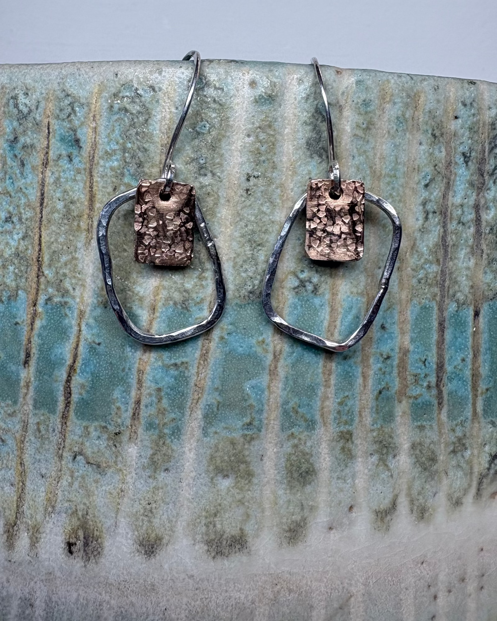 Pair of silver hoop earrings with bronze textured squares on a ceramic surface
