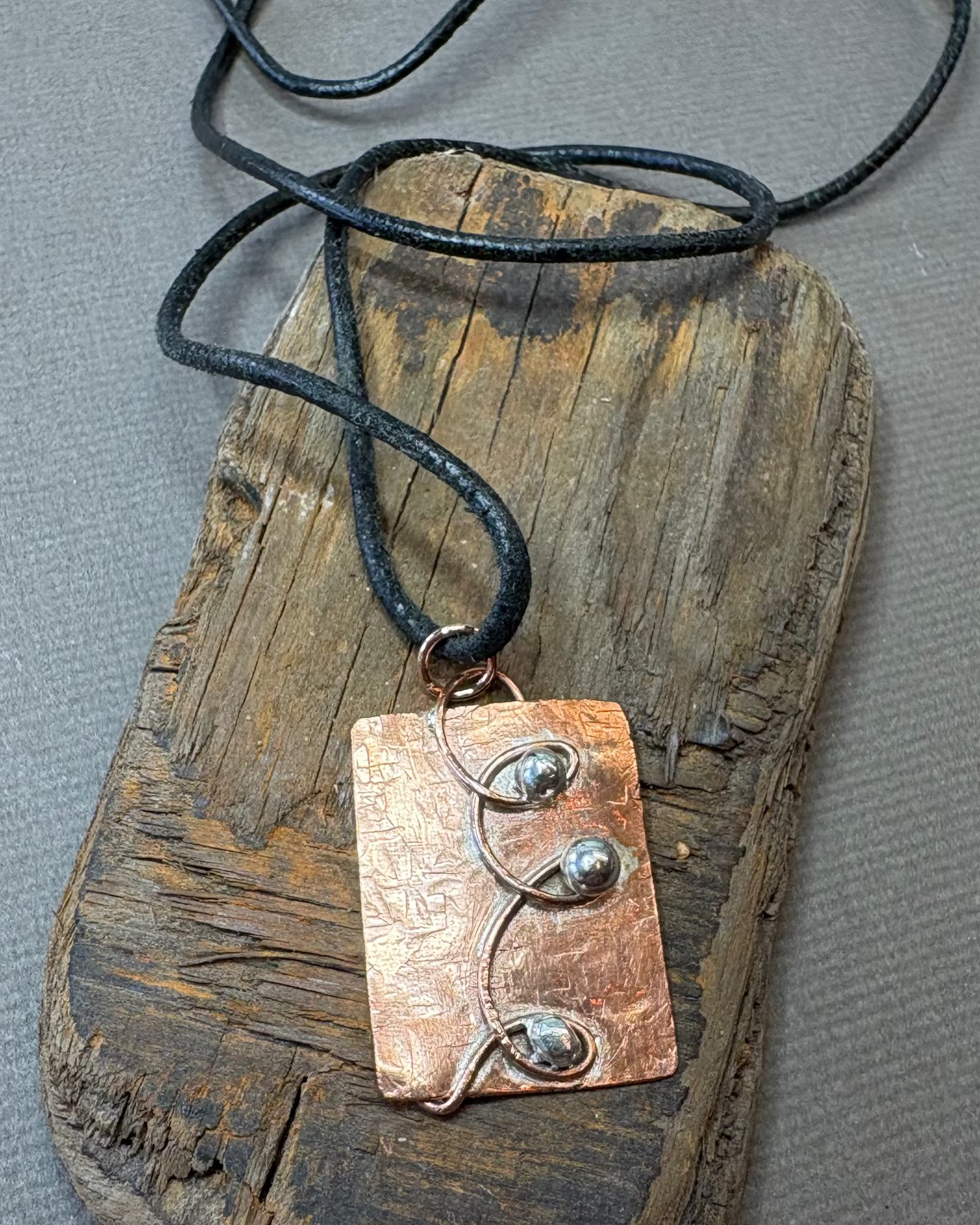 Copper pendant with silver accents with a black cord, placed on a piece of driftwood