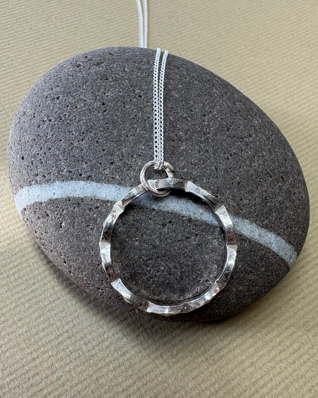 Silver necklace with a circular wavy pendant on a pebble surface