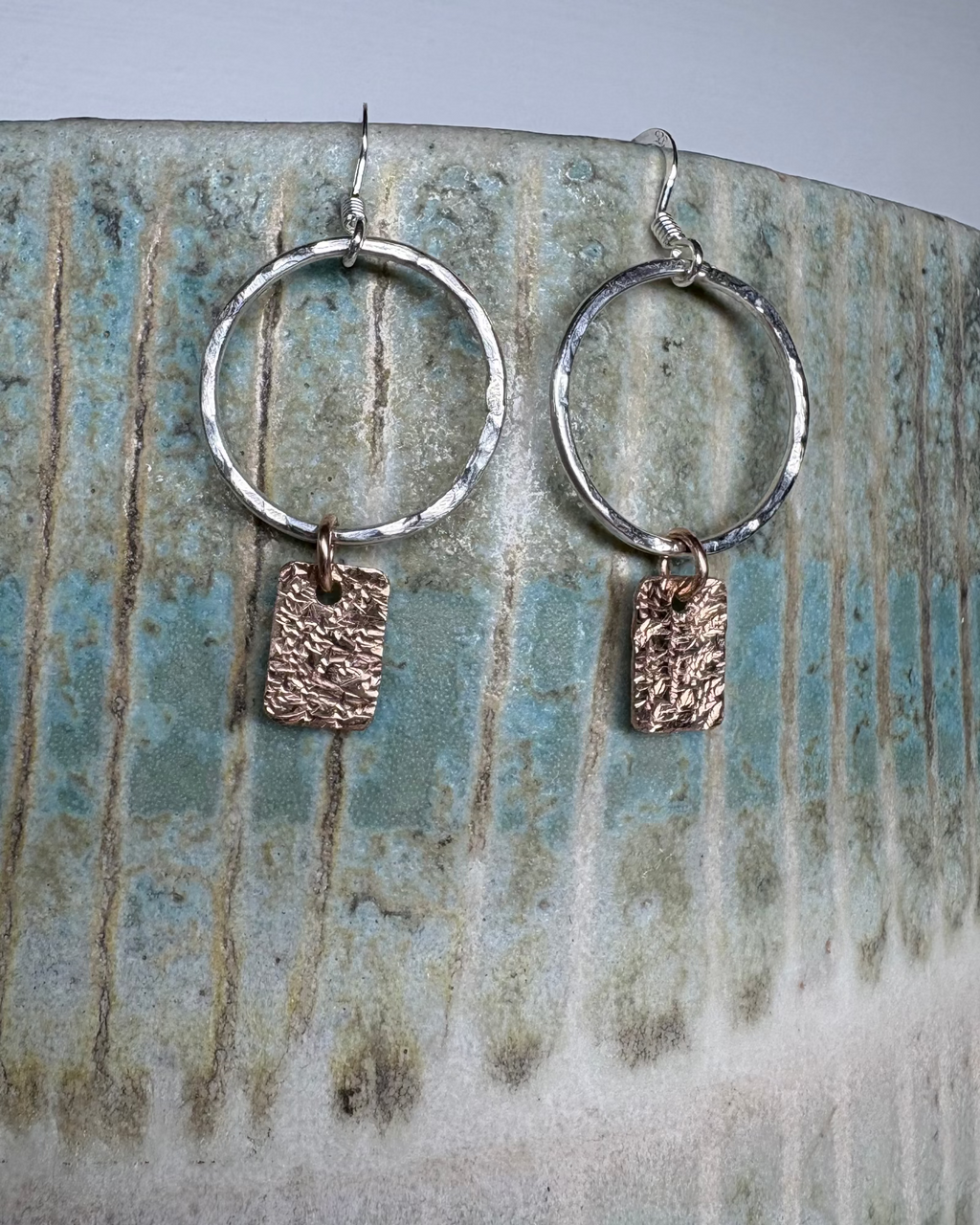 Silver hammered hoop earrings with bronze textured charms on a ceramic surface