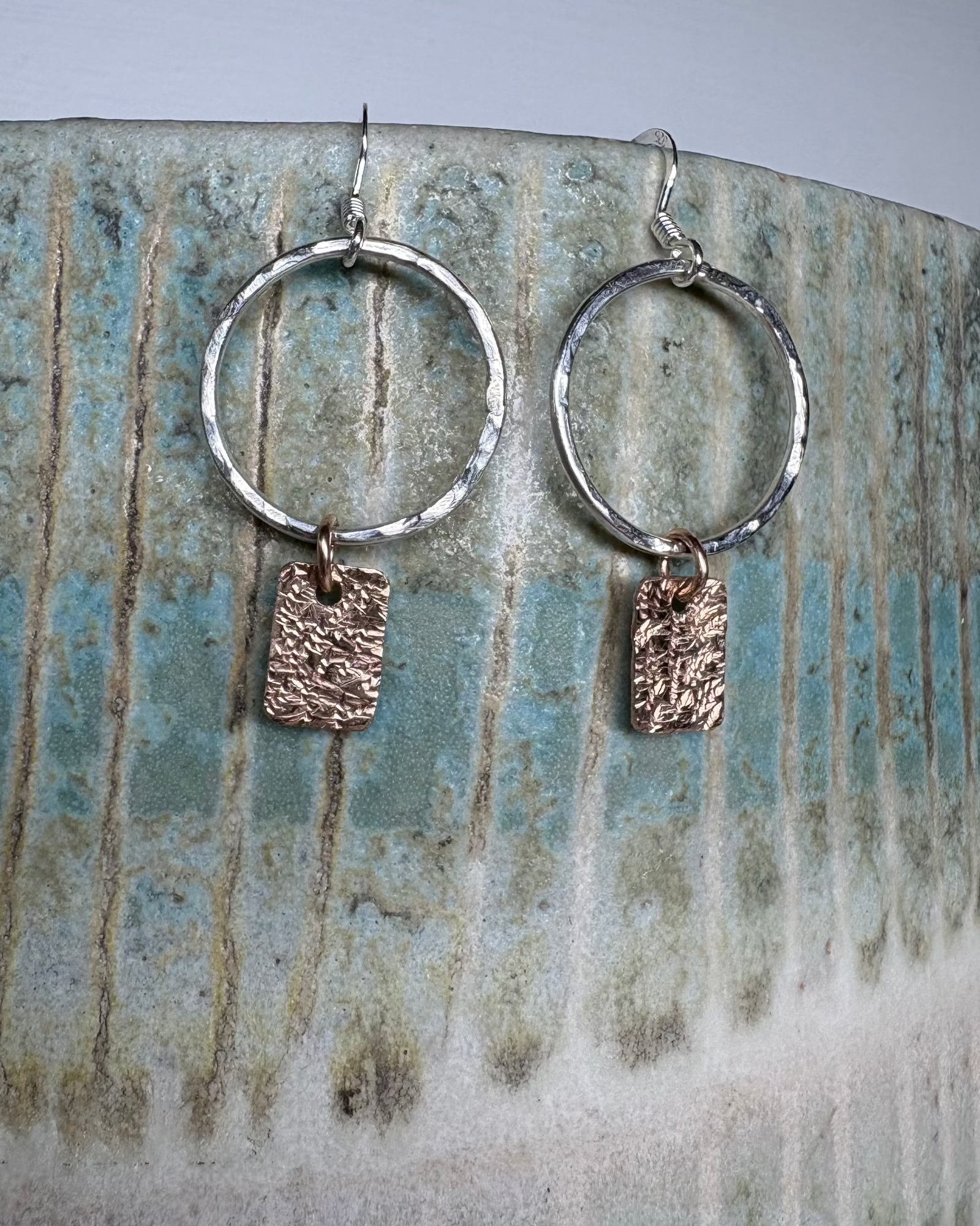 Silver hammered hoop earrings with bronze textured charms on a ceramic surface