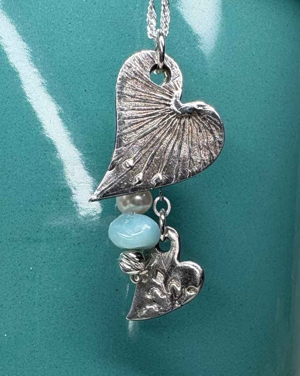 Necklace with heart-shaped charms with an apatite bead and freeform pearl hanging on a silver chain against teal background