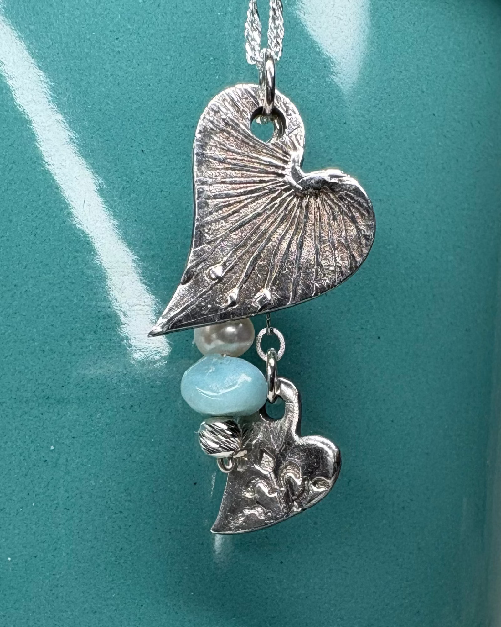 Necklace with heart-shaped charms with an apatite bead and freeform pearl hanging on a silver chain against teal background