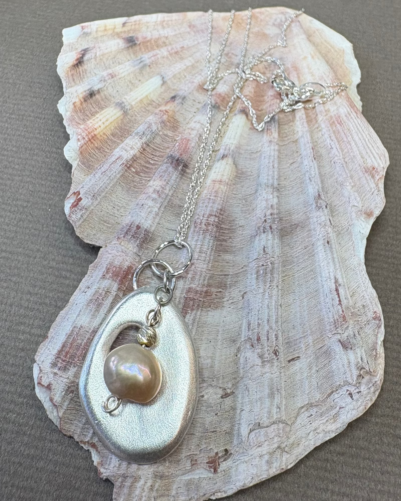 Holey shell silver form pendant with hanging natural pearl pictured on a shell