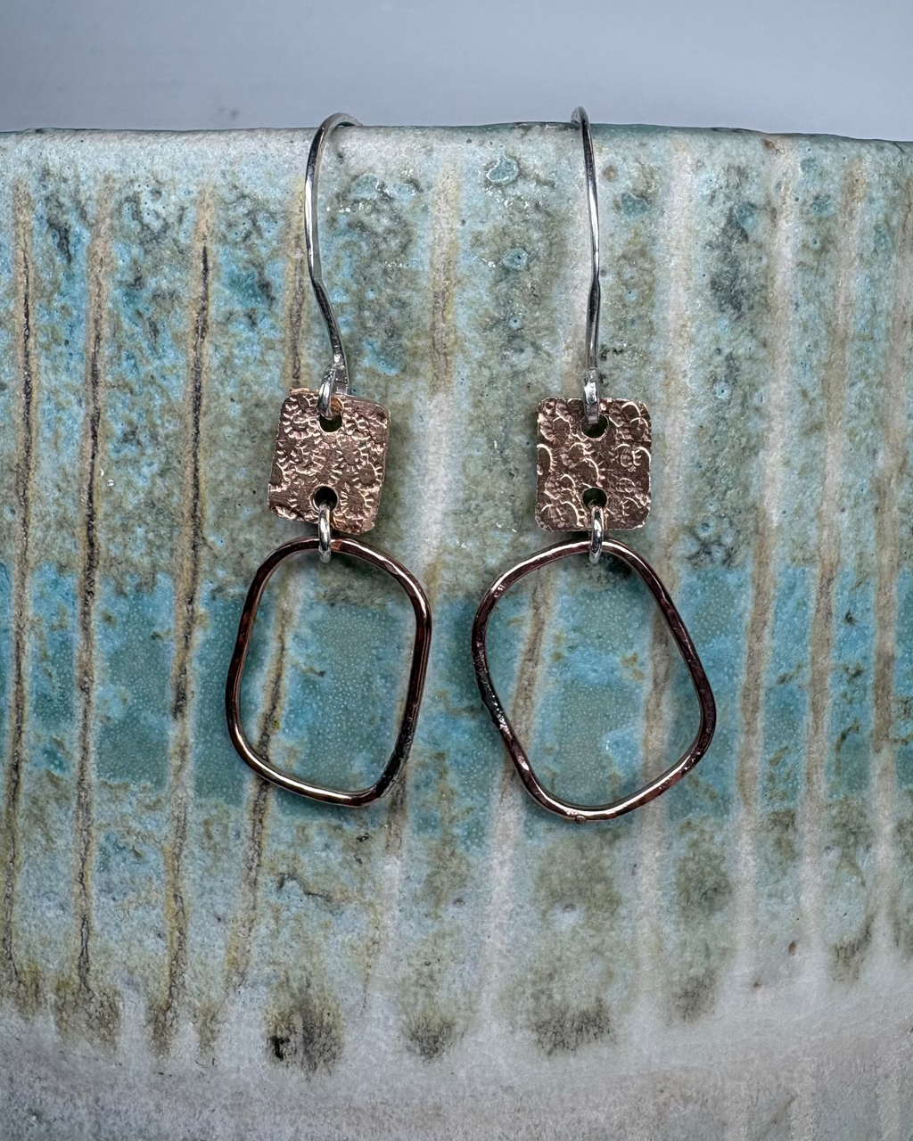 Pair of hammered bronze hoop and textured charm earrings on a ceramic surface