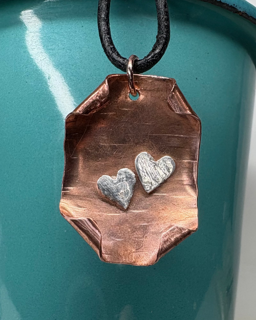 Copper and silver heart pendant with curled corner design with a black leather cord pictured on a teal container