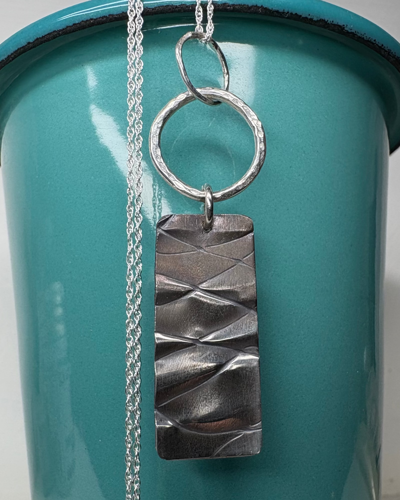 Necklace with a textured rectangle under two rings with a silver chain on a teal background