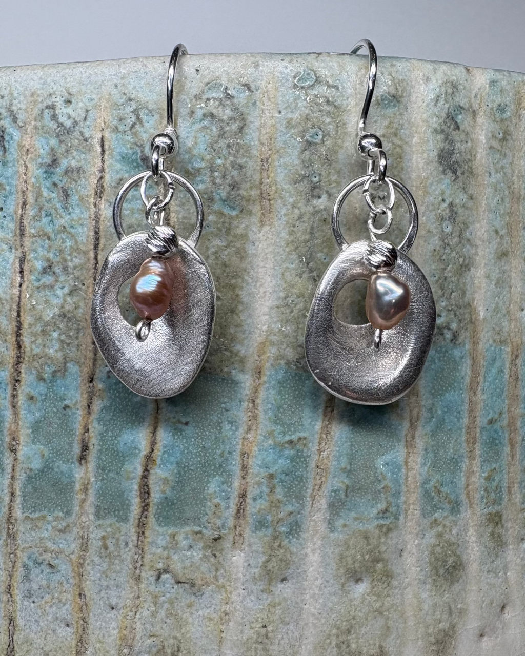 Pair of silver earrings with a central pearl on a textured ceramic surface.