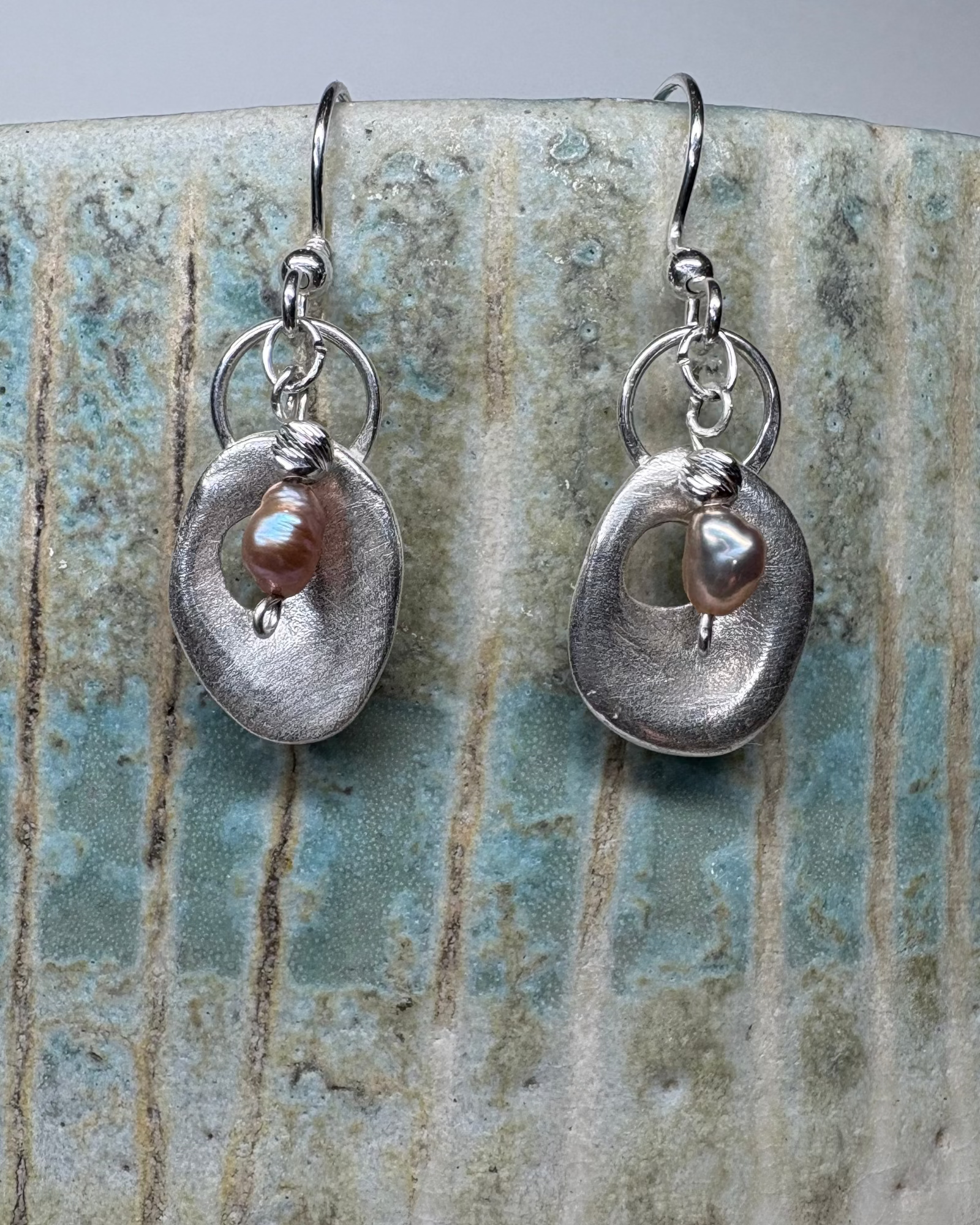 Pair of silver earrings with a central pearl on a textured ceramic surface.