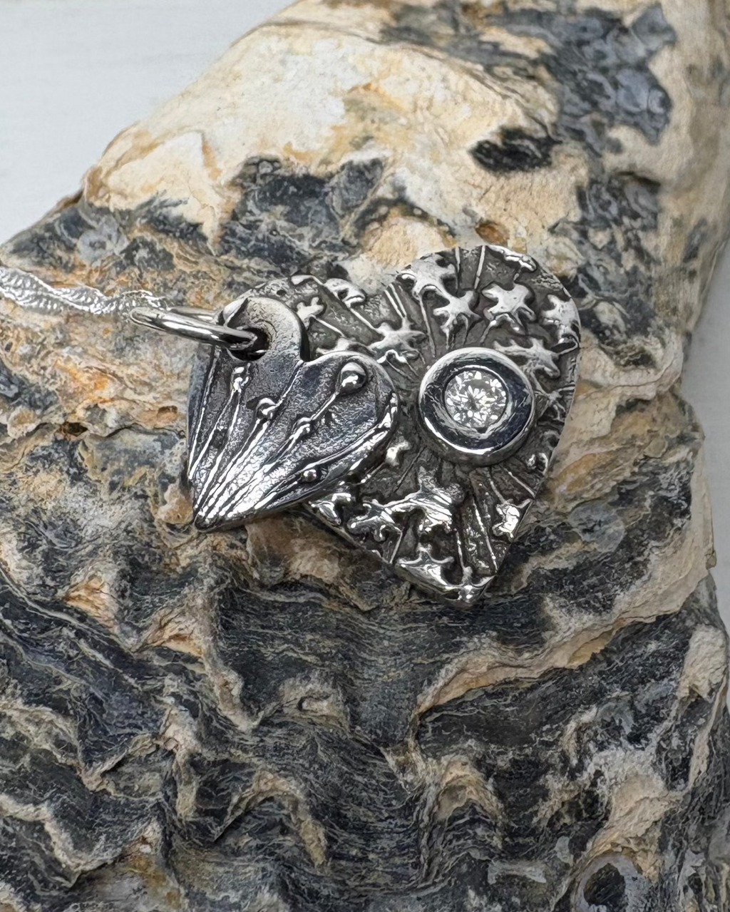 Silver oxidised double heart pendant with textured seed head designs with a white cubic zirconia set on larger heart with silver chain pictured on a textured shell